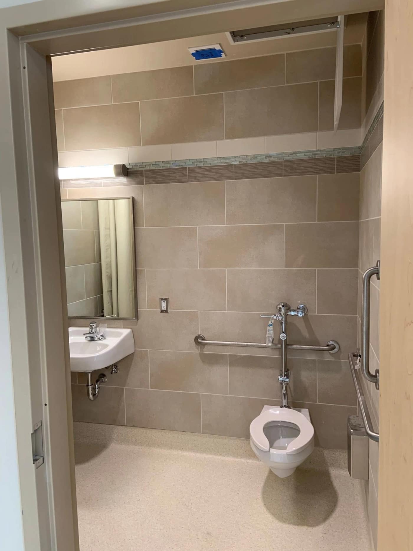 An accessible bathroom in tan large-format tile with a glass mosaic band at the Bethel hospital project